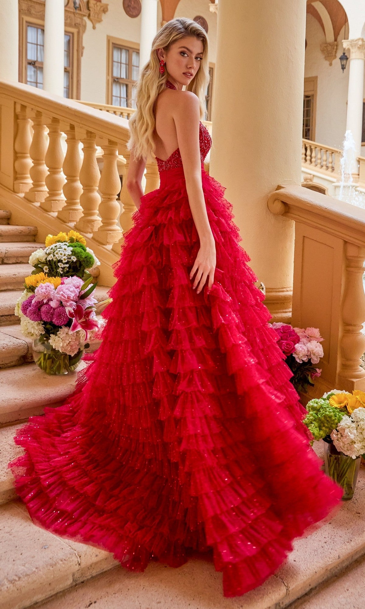 A woman wearing the Ava Presley 47258 long prom dress, a strapless red layered ruffled ball gown, stands on a staircase adorned with flowers in an elegant building with columns and arches.